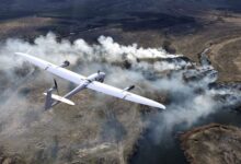 A white Vector reconnaissance drone is seen flying a gray landscape. The drone has the shape of a typical airplane, with a slimmer body and wings. Some areas of the field below have smoke billowing out from what appears to be burning grass. Trees with little to no leaves are dotted around the area.