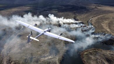 A white Vector reconnaissance drone is seen flying a gray landscape. The drone has the shape of a typical airplane, with a slimmer body and wings. Some areas of the field below have smoke billowing out from what appears to be burning grass. Trees with little to no leaves are dotted around the area.