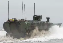 An Amphibious Combat Vehicle is seen trudging on ocean waters. The vehicle has four antennae on top of it, all pointing straight up. It's painted in camouflage. A misty mix of white and muddy brown water surrounds it as it traverses the waters.