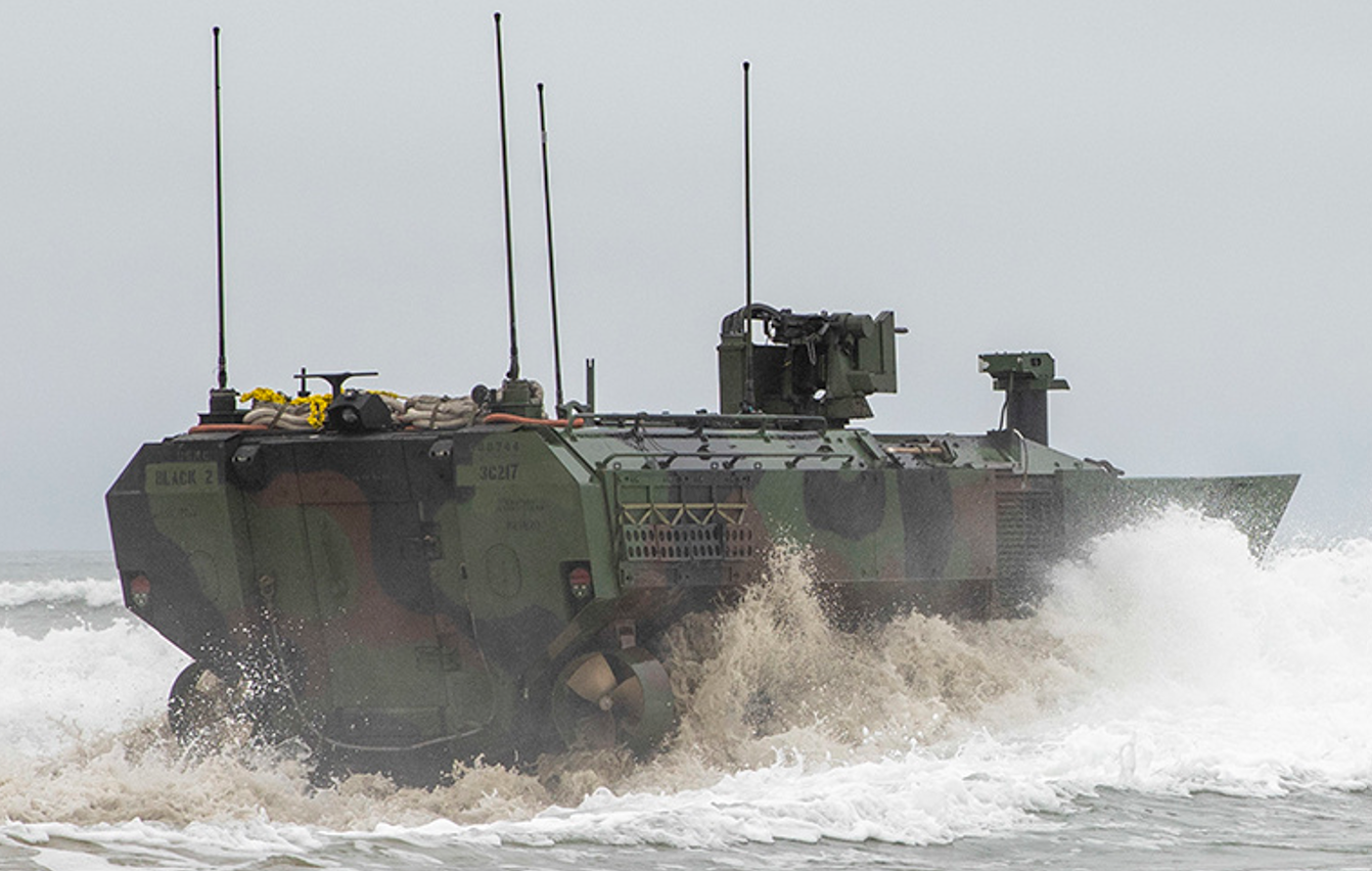 An Amphibious Combat Vehicle is seen trudging on ocean waters. The vehicle has four antennae on top of it, all pointing straight up. It's painted in camouflage. A misty mix of white and muddy brown water surrounds it as it traverses the waters.
