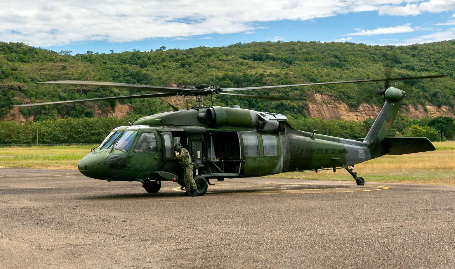 A Colombian Army Black Hawk helicopter is seen grounded in a large open cemented field. The helicopter is painted various hues of dark green as camouflage. A soldier in camouflage is seen standing by the cockpit. "EJERCITO DE COLOMBIA" is seen painted in black on the aircraft's tail boom. The background is a mountain covered by a lush green forest, and a bright blue sky with big white clouds.