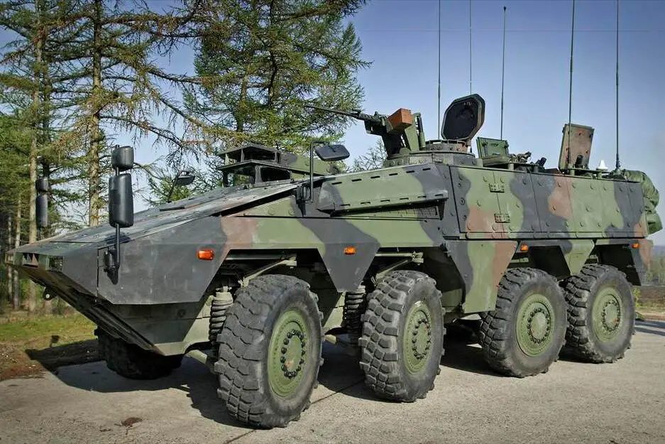 A Boxer armored fighting vehicle is seen parked in a cemented area in front of a green, tree-filled field. The eight-wheel armored vehicle is painted in green, brown, and black camouflage. A mounted gun by its hatch above is seen pointing forward. The background is a clear blue sky.