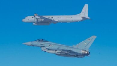 A German Eurofighter flies below another aircraft. Both planes are painted gray, with the Eurofighter having a darker tone. The background is a clear blue sky.