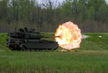 The US Army's M10 Booker during a live fire demonstration. The armored vehicle is seen firing a round from its 105 mm gun. A ball of red-orange fire is seen coming out of the weapon, with the ammunition flying through it. The vehicle is colored dark green. The location where the demonstration is happening is a green field with a forested area in the background.