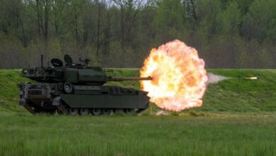 The US Army's M10 Booker during a live fire demonstration. The armored vehicle is seen firing a round from its 105 mm gun. A ball of red-orange fire is seen coming out of the weapon, with the ammunition flying through it. The vehicle is colored dark green. The location where the demonstration is happening is a green field with a forested area in the background.