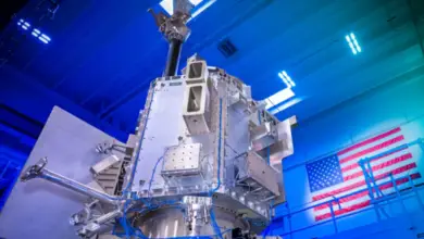 BAE Systems' Weather System Follow-on Microwave (WSF-M) satellite. The satellite appears to have a chrome silver finish, with blue wiring connecting some of its parts together. The background shows that it's displayed in a warehouse-like building with blue overhead lights. In the back, a Unites States flag can be seen displayed on a wall.
