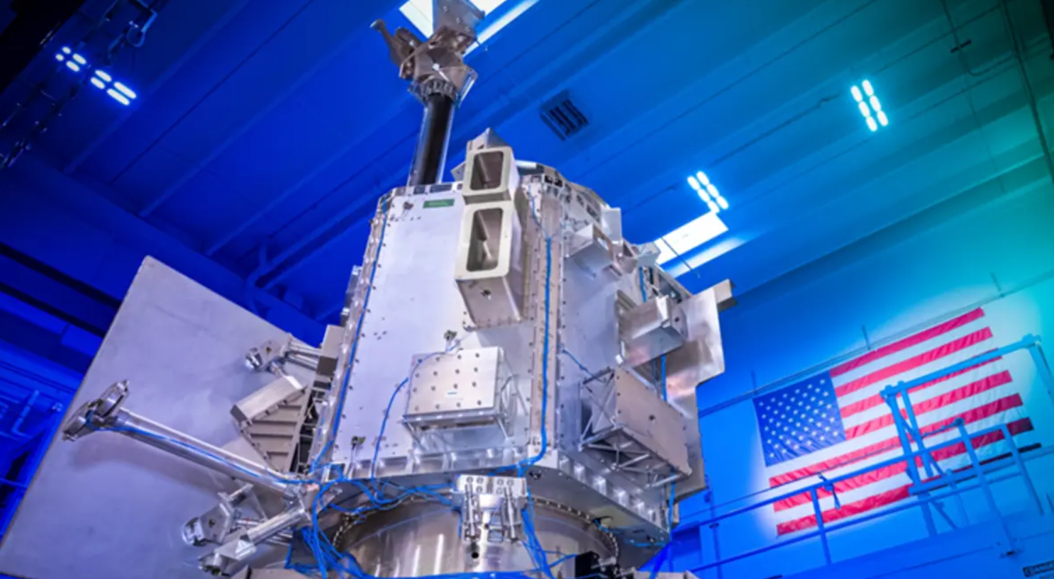BAE Systems' Weather System Follow-on Microwave (WSF-M) satellite. The satellite appears to have a chrome silver finish, with blue wiring connecting some of its parts together. The background shows that it's displayed in a warehouse-like building with blue overhead lights. In the back, a Unites States flag can be seen displayed on a wall.