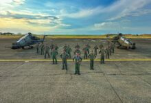 21 personnel from the Philippine Air Force pose in attention between two T129 ATAK helicopters in what appears to be a large landing area for helicopters. The soldiers are all wearing dark green fatigues and blue side caps. The helicopters are painted dark green. The background is a vast blue sky with some clouds, and colors of the sunset beginning to show in the far left side of the image.