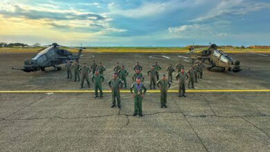 21 personnel from the Philippine Air Force pose in attention between two T129 ATAK helicopters in what appears to be a large landing area for helicopters. The soldiers are all wearing dark green fatigues and blue side caps. The helicopters are painted dark green. The background is a vast blue sky with some clouds, and colors of the sunset beginning to show in the far left side of the image.