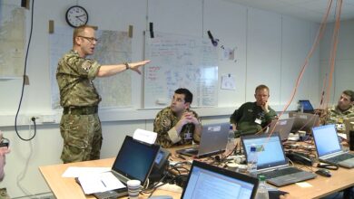 Three uniformed military officials are seen in a meeting, with one official standing up and raising his right arm to gesture to the group and the other two listening while sitting at a table. The room they are in appears to be an office, with maps, graphs, and various illustrations attached to the whiteboard and the wall in the background. Laptops and other electronic devices are propped up on the table.