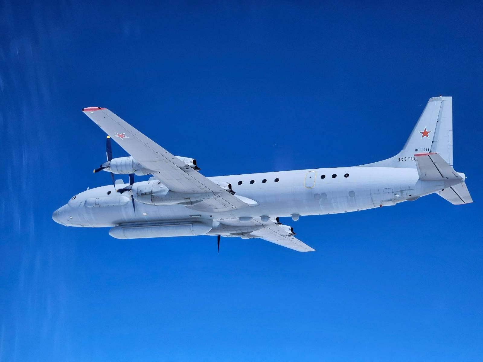 A gray Russian Il-20 aircraft, with tail ID RF-93611, is seen flying in clear blue skies. The image is taken a little below the left side of the aircraft. The Il-20 has two propellers attached on each of its wings.