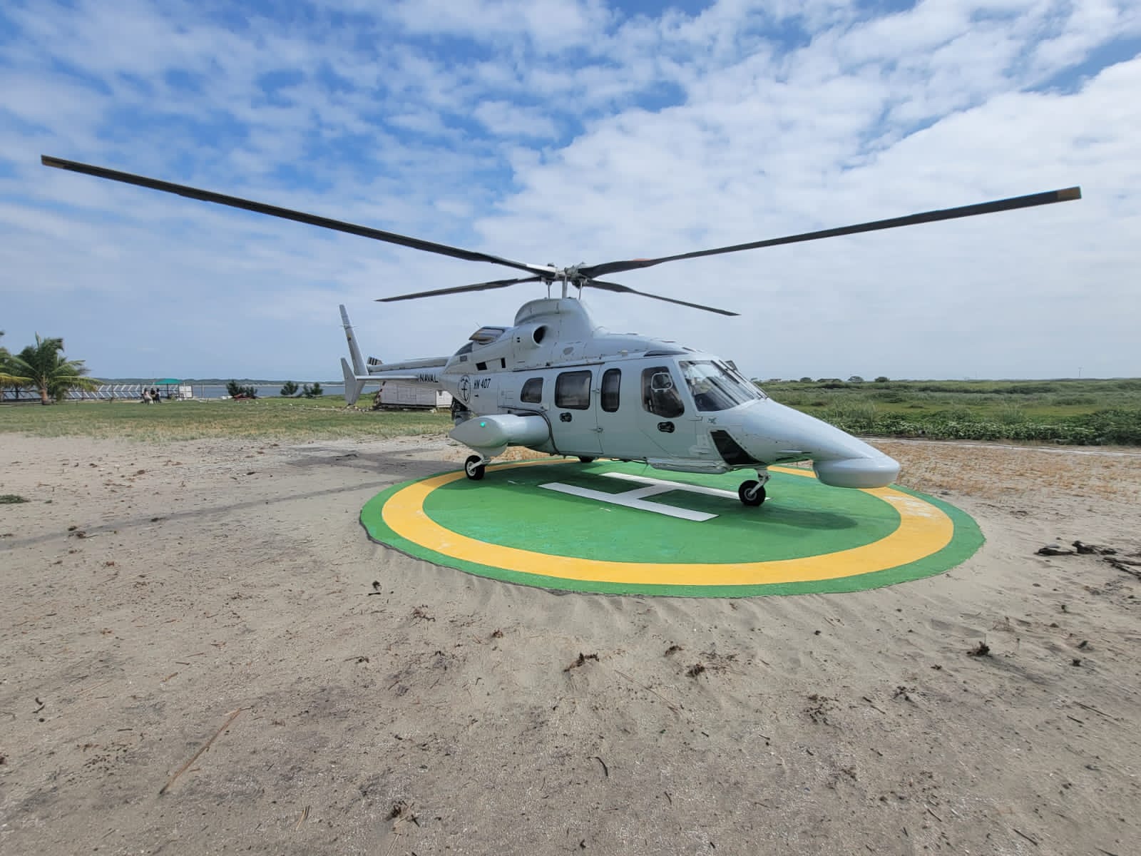 The Ecuadorian Navy's Bell 430 aircraft with the ID HN-407 is seen on a helipad in the middle of a sandy area. The helicopter is painted white, with "HN-407" written on the right side of its body in black. On the back, green grass can be seen stretching all the way to the horizon. A body of water peeks a little bit farther. The sky is blue with lots of thin white clouds.