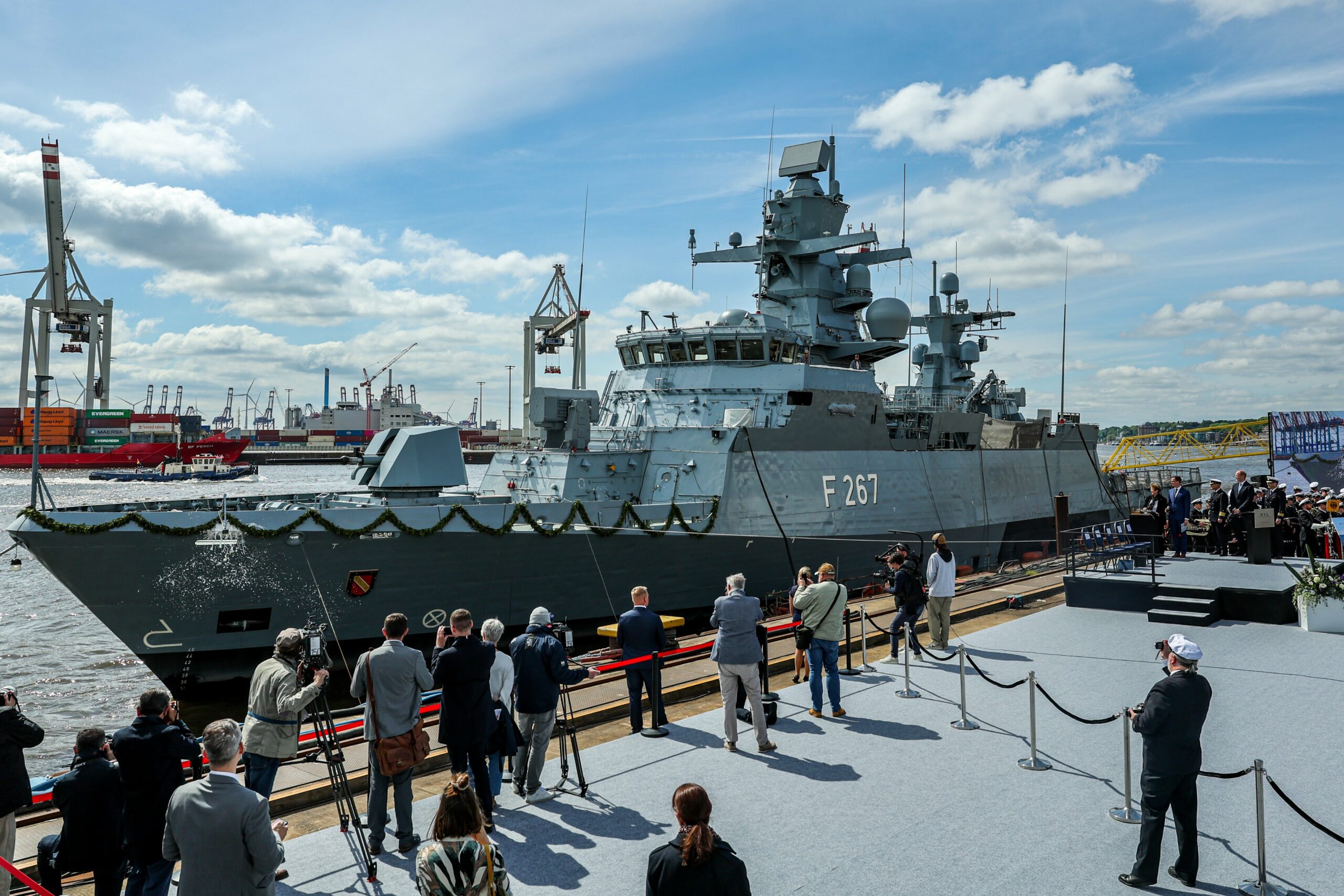 The Karlsruhe, a German Navy corvette, is seen docked in a pier during its christening ceremony. The gray ship's deck railings are decorated with a ribbon made of leaves. The middle of its hull has "F267" painted on it in white. A group of journalists, photographers, and navy officers and personnel are positioned on the dock, observing the corvette. The background is a clear blue sky with some light white clouds.