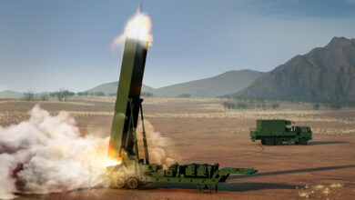 A Long Range Hypersonic Weapon (LRHW) system is seen firing off a missile upwards in the middle of a brown plains area. The system is attached to a six-wheel hydraulic launching system that attaches to a six-wheel truck. The truck is seen a little farther back, on the right side of the image. Both the vehicle and the weapon system is painted green. The background is a scenic view of a mountain range, with a grayish-blue sky occupying the rest of the space.