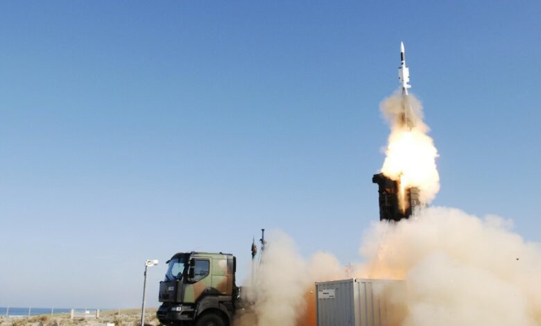 A SAMP/T air defense system is seen firing off a ballistic missile from the back of the military truck. Smoke and bright white-red fire can be seen jutting out of the missile as it flies straight upward. The background is a clear blue sky.