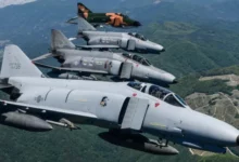 Four F-4E Phantom jet planes are seen flying over a green landscape in South Korea. Three of the planes are painted gray, while the one in the far left is painted camouflage green and orange.