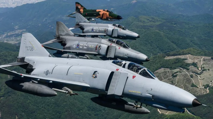 Four F-4E Phantom jet planes are seen flying over a green landscape in South Korea. Three of the planes are painted gray, while the one in the far left is painted camouflage green and orange.