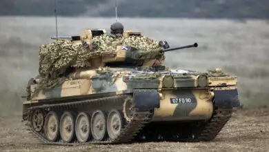 An FV107 Scimitar armored tracked reconnaissance vehicle fleet is seen in the middle of a plains. The vehicle is painted camouflage brown and green. A soldier wearing protective goggles and a green helmet is seen peeking out from the vehicle's hatch. The background is out of focus but appears to be a forested area.