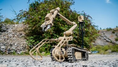 An L3Harris T4 explosive ordnance disposal robot is displayed on a flat, rocky area with bushes in the background. The robot has tracked wheels similar to a tank, two arms resesembling a mantis' raptorial legs, and a crane mechanism with a mechanical claw at the end. The unit is painted brown-yellow.