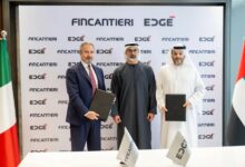 Fincantieri CEO and Managing Director Pierroberto Folgiero, Abu Dhabi crown prince Khaled bin Mohamed Al Nahyan, and EDGE Group chairman Faisal Al Bannai stand to take a photo op during a signing ceremony. Folgiero and Al Bannai hold leather dosiers, which may be holding the signed documents. Italy's flag is seen displayed on the left, and UAE's is on the right. On a white table in front of them, two flags printed with Fincantieri and EDGE's logos are also propped up. Behind them is a white screen with the companies' logos printed in a repeating pattern.