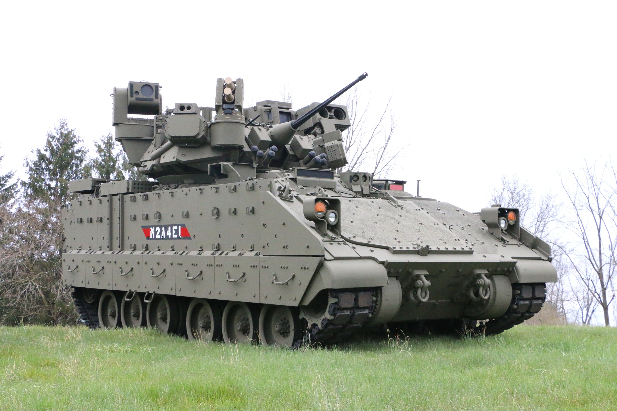 A Bradley M2A4E1 Fighting Vehicle is seen stationed at a grassy area with thin trees in the back. The vehicle is a tracked armored unit that is painted grayish green. Its turret points northeast, and its body is facing to the right side of the image. Its model name is painted in white on its right side, with a gray and red background.