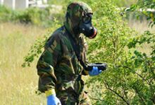 A soldier wearing a gas mask and camouflage fatigues is seen holding a ChemProX handheld chemical detector in a grassy field.