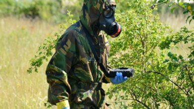 A soldier wearing a gas mask and camouflage fatigues is seen holding a ChemProX handheld chemical detector in a grassy field.