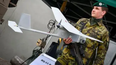 A soldier wearing camouflage fatigues and a green beret is seen holding an unmanned aerial vehicle with one hand. The small gray drone has a simple shape similar to that of high-wing airplanes.