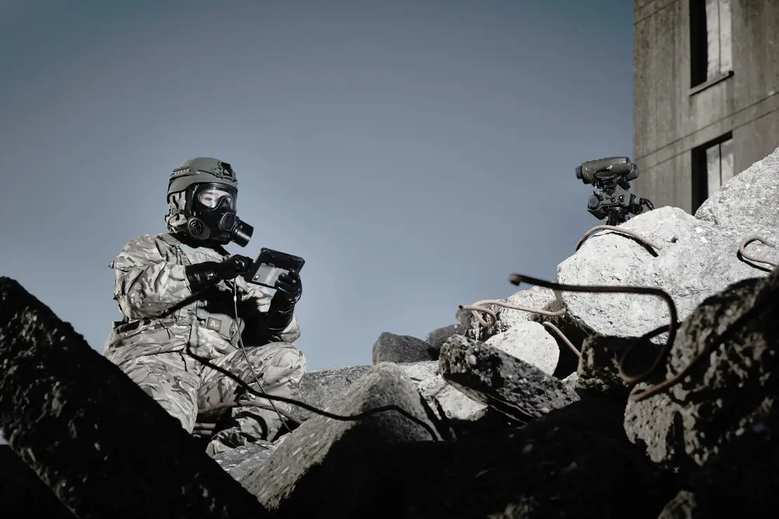 A soldier wearing camouflage fatigues and a General Service Respirator mask is seen using a wired device the size of a small tablet in a pile of rubble.