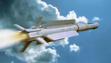 An IRIS-T missile is seen flying in a cloudy blue sky. It juts out smoke and red and white flames as it propels upward. The missile is sleek, painted gray, and has four fins attached to its bottom.