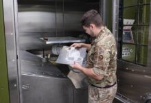 A British Army soldier examines the 3D printer's output. He holds a sheet of metal with the printed white product attached to it. It is likely a spare part for a military equipment. The soldier is wearing brown camo fatigues. The 3D printer is approximately the size of a photo booth, with riveted sheet metal finishings and some panels painted moss green.
