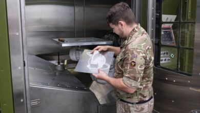 A British Army soldier examines the 3D printer's output. He holds a sheet of metal with the printed white product attached to it. It is likely a spare part for a military equipment. The soldier is wearing brown camo fatigues. The 3D printer is approximately the size of a photo booth, with riveted sheet metal finishings and some panels painted moss green.